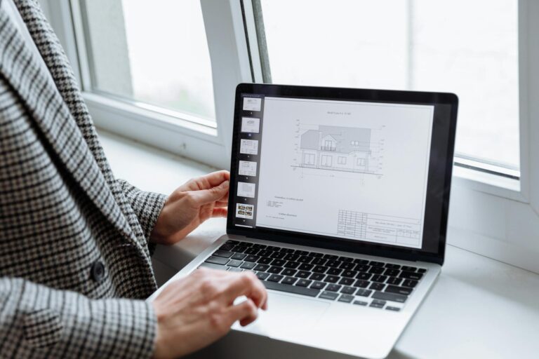 Person in plaid blazer reviewing architectural blueprints on laptop by a window.